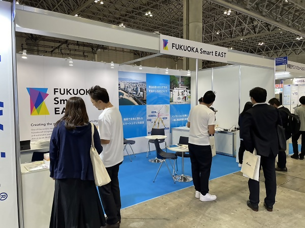 Fukuoka Smart Eastブースの風景画像。立ち寄ってくださった方にスタッフが説明しています。